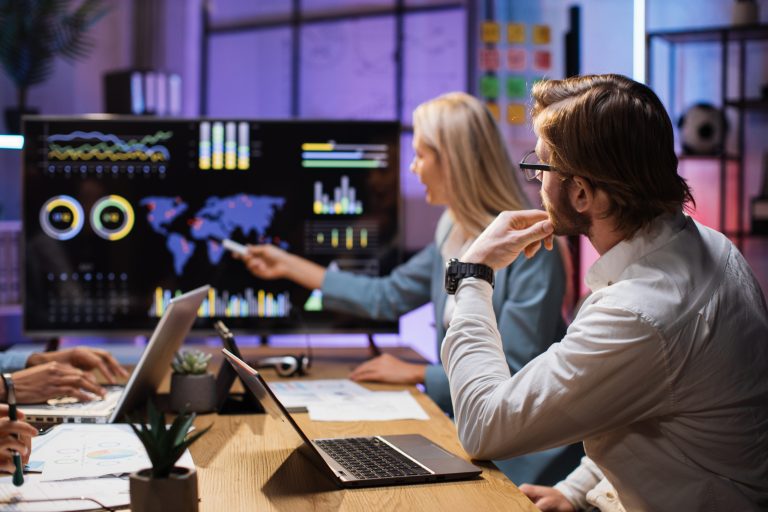 Group of competent financiers having working meeting at office during evening time. Partners examining report of company. Focus on big computer monitor with statistics graphs.